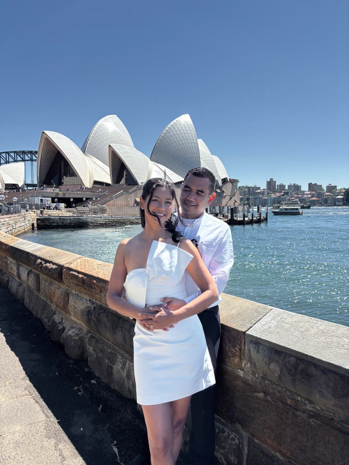 Pilar y Juan abrazados y sonriendo junto a la bahía, con la Ópera de Sídney al fondo